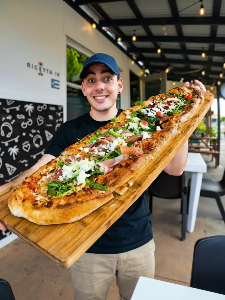 Un visitante feliz en La Ricetta con una pizza alargada gigante de prosciutto, arúgula, queso de cabra y tomates cherry